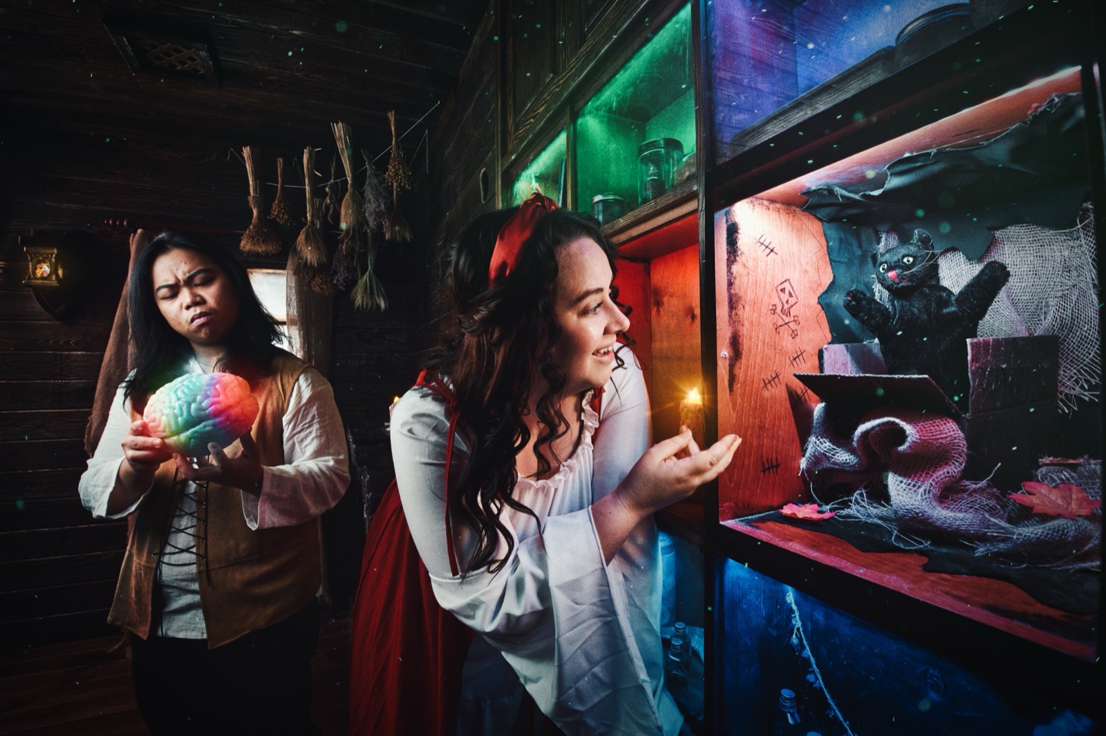 In the witch's cabin of Malediction, two women in costume: one holds a glowing orb, one reaches toward a black cat